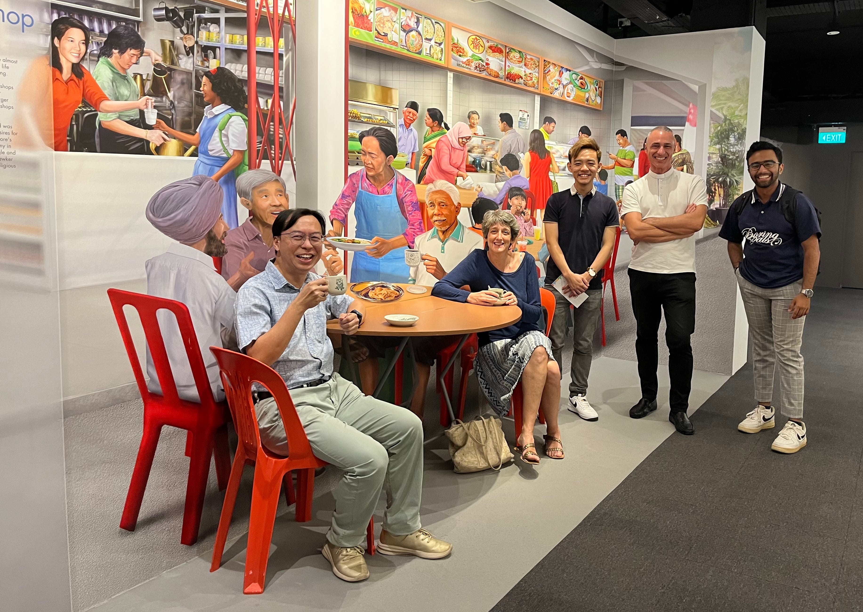 Five people pose at a museum exhibit featuring a large mural of a busy hawker food centre scene.